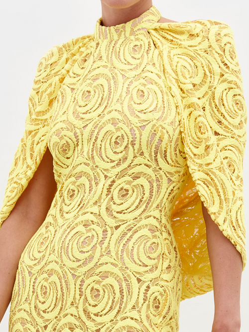 Person wearing a Salvina Dress Tuscany Yellow, adorned with a high neckline and cape-like sleeves, holding a pearl bracelet on their wrist, eagerly awaiting the pre-order ship date for 2025.