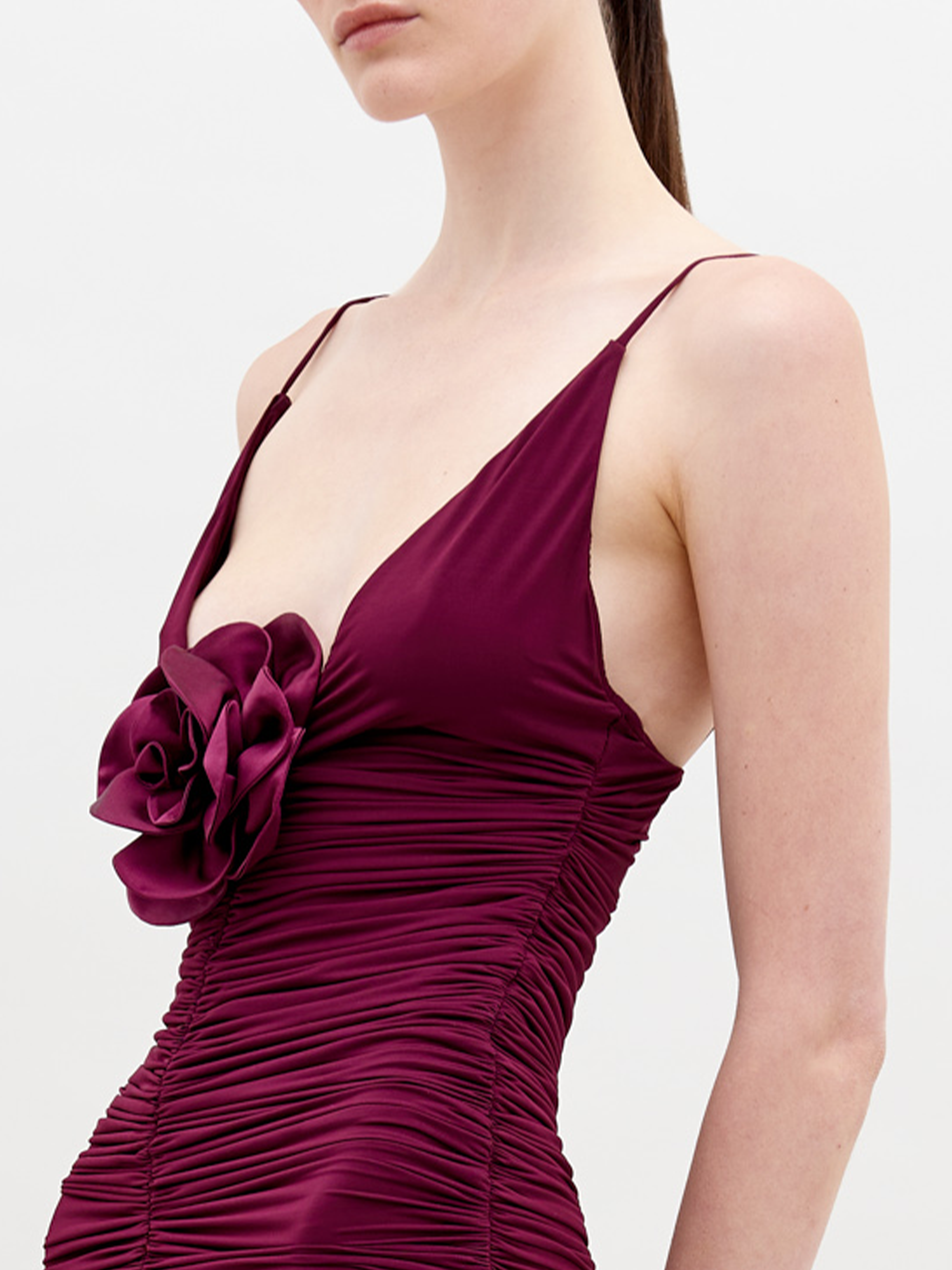 A person is wearing the SVT DESIGNS Sana Dress, a sleeveless deep burgundy maxi with thin straps and a prominent 3D floral appliqué rosette on the chest. It has ruched fabric from bust to waist against a plain white background.
