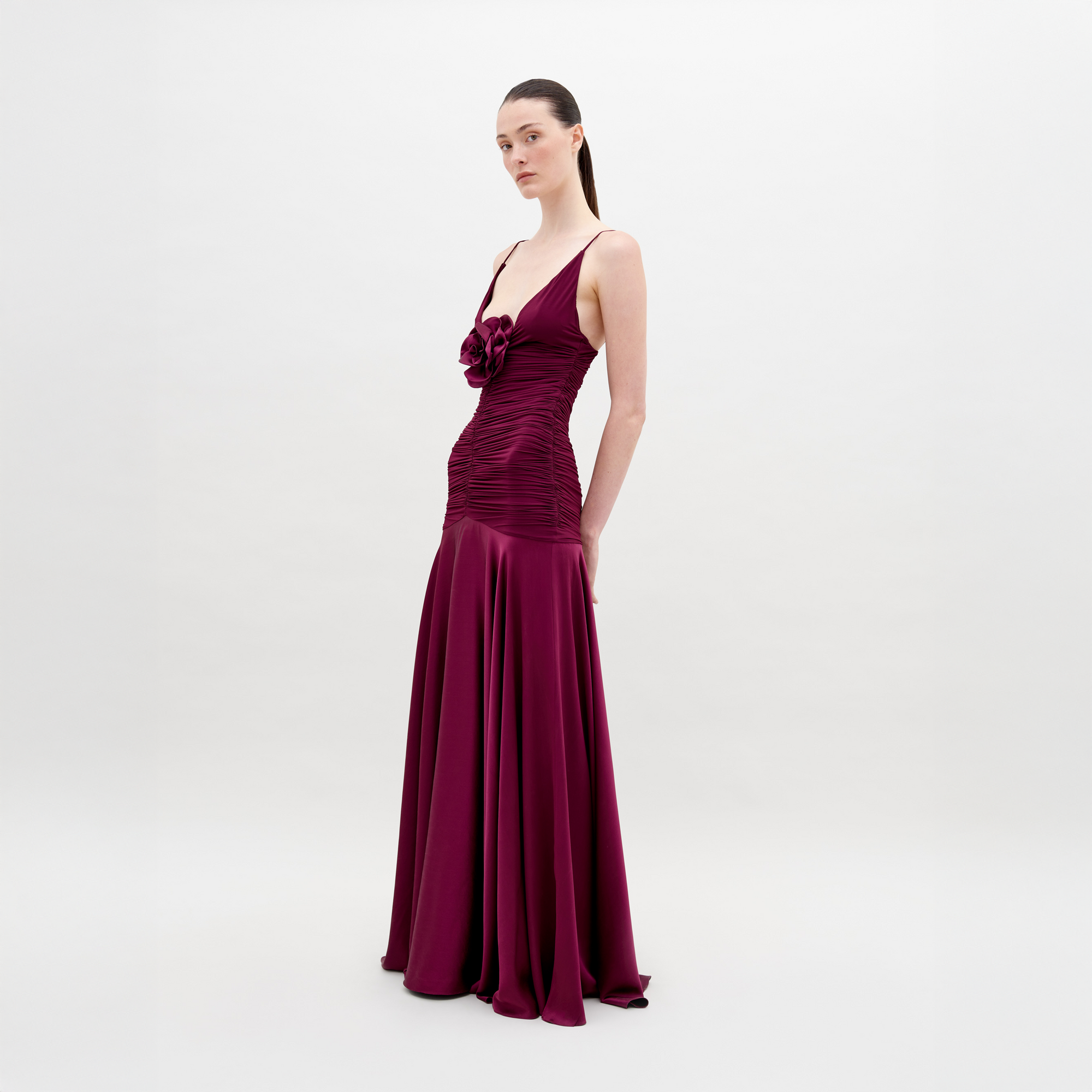Against a white background, a person wears the SVT DESIGNS Sana Dress, a floor-length maxi in deep magenta. It showcases an Italian textile with a fitted, ruched bodice and 3D floral appliqué on the chest. Their hair is pulled back as they glance slightly aside.
