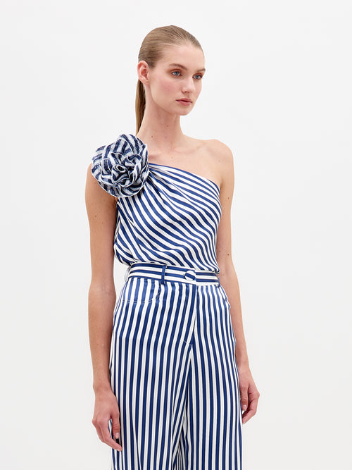 Someone wears the Sebastiana Blouse by SVT DESIGNS, a blue and white striped silk one-shoulder jumpsuit with a floral appliqué on the shoulder. It features vertical stripes and a belted waist for an elegant silhouette against a plain white background.