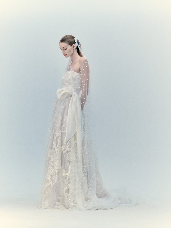 A woman in a Selman Dress Pearl with a flowing veil poses against a light blue background.