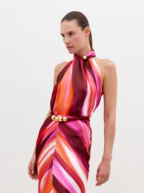 A woman stands against a plain background, wearing a halter-neck dress with a multicolored pattern of pink, orange, and red hues, accessorized with a gold belt buckle. Pre-order the Shela Blouse Magenta Abstract Waves now to ensure delivery by November 15th, 2024.