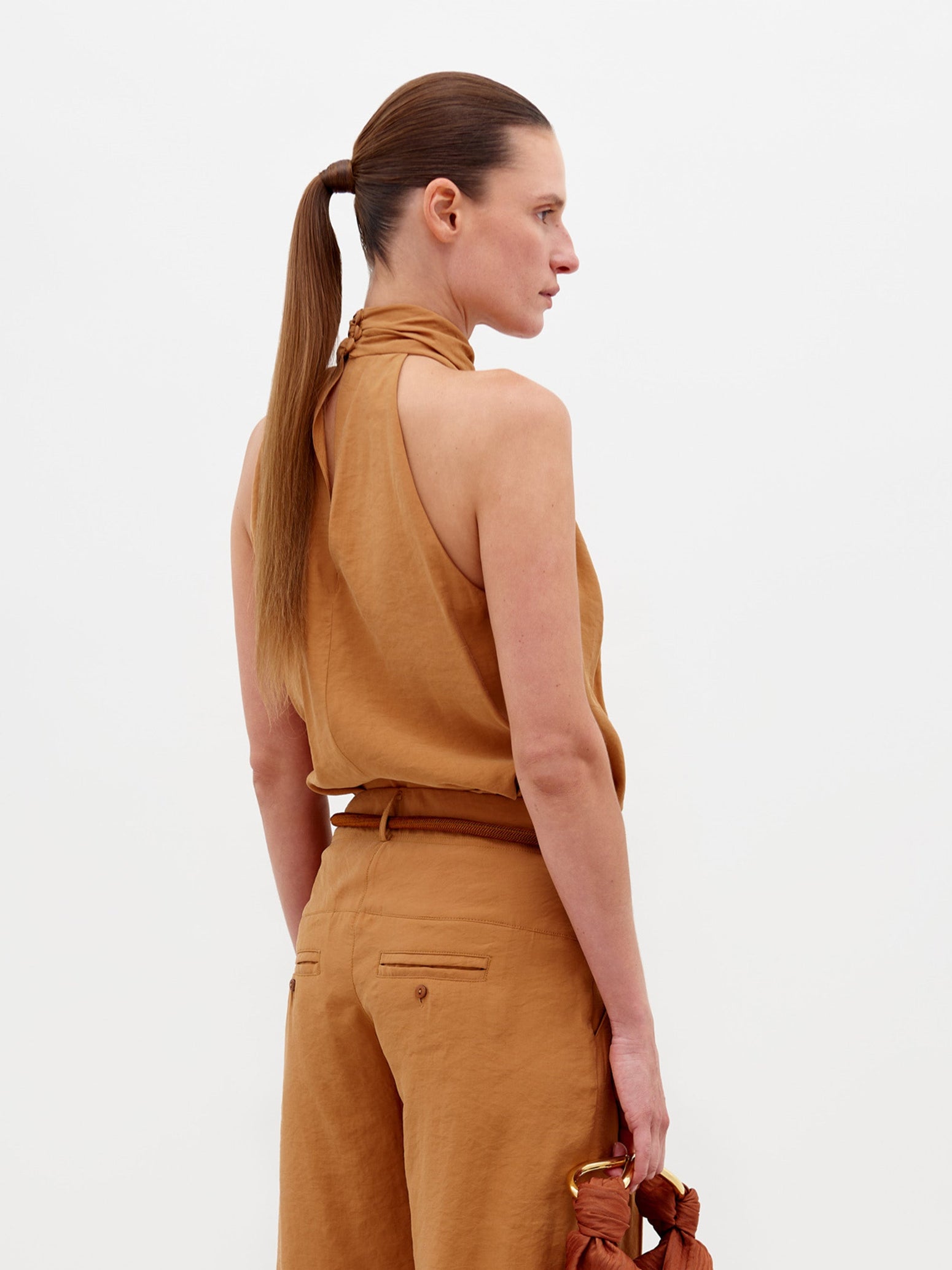 A person wearing the Shela Blouse Camel sleeveless high-neck top with matching wide-leg trousers, accessorized with a belt and holding a small object, stands against a plain white background. Pre-order now to ensure it ships by February 15th, 2025.