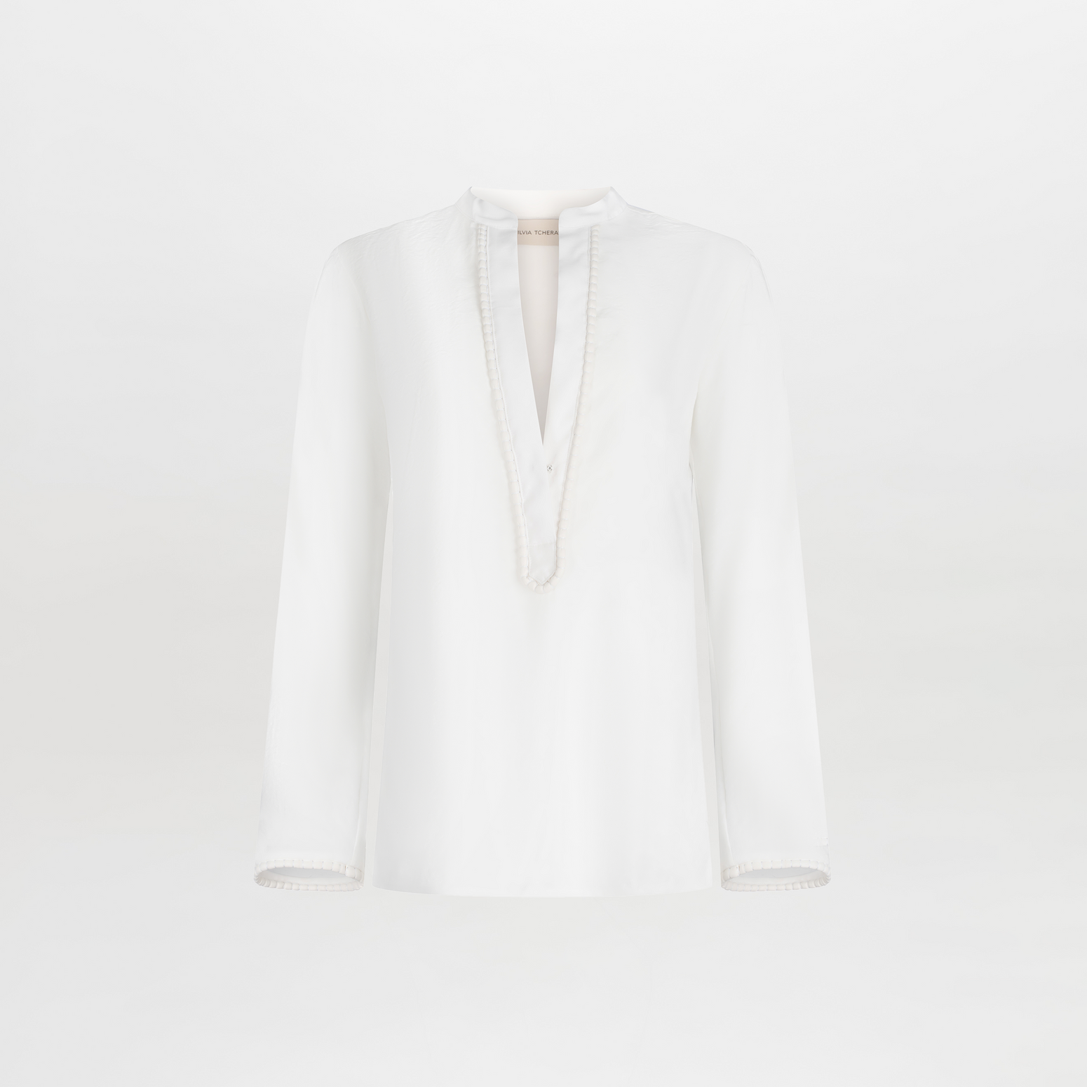 The Shera Blouse by SVT DESIGNS is a white, long-sleeve top with a V neckline, featuring subtle fringe lace on the deep cut and cuffs. It has a loose silhouette for a relaxed fit and is displayed against a plain white background.