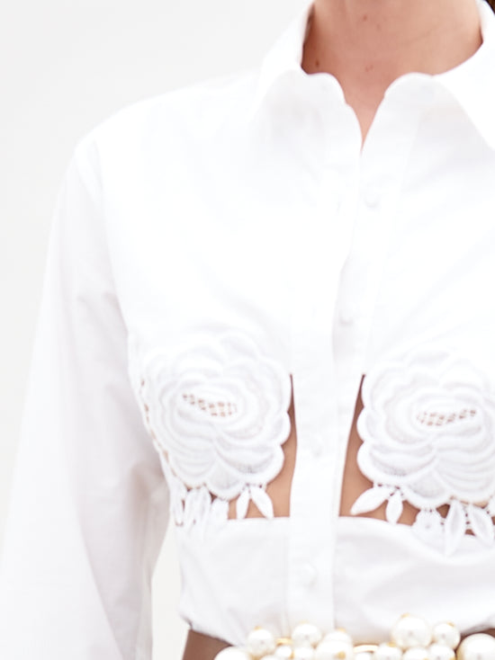 A person is wearing the Sigrid Blouse by SVT DESIGNS, a white cotton shirt featuring intricate floral lace appliqué on the chest and elegant cut-out sections. The blouse is highlighted with a belt adorned with large pearl-like ornaments, emphasizing its exquisite details.