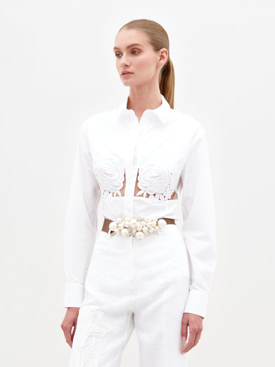 A person wears the Sigrid Blouse by SVT DESIGNS, a white cotton blouse with floral appliqué and cut-out details, paired with high-waisted white trousers. Their long hair is tied back as they stand elegantly against a plain white background.