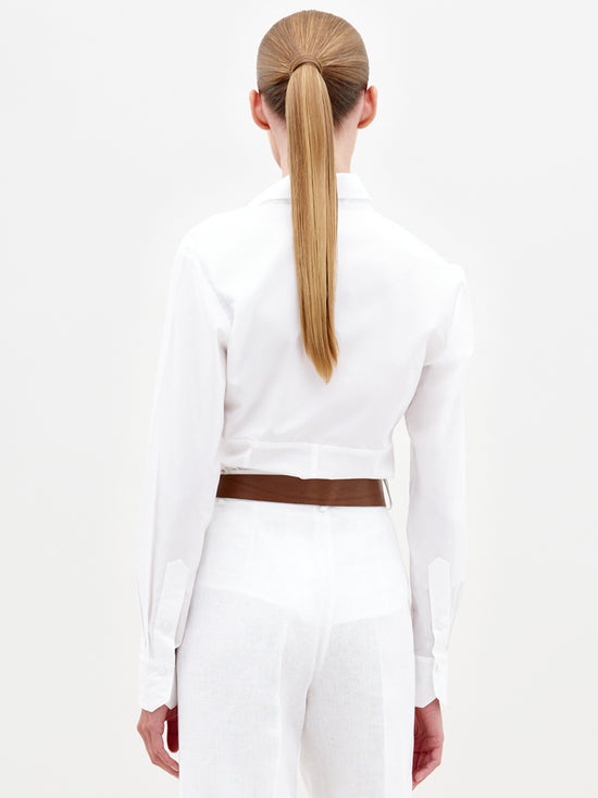 With long blonde hair in a ponytail, the person is wearing the Sigrid Blouse and white pants, highlighted by a brown belt. Crafted by SVT DESIGNS with subtle Italian-inspired details, their chic ensemble stands out against the minimalist backdrop.