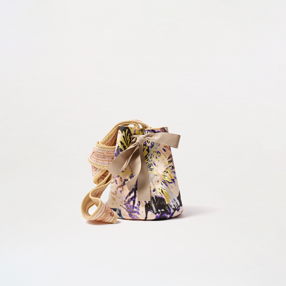 The Mochila Laaput Small is a colorful bucket bag with a floral pattern, decorative bow, beige ribbon tie, and woven shoulder strap, made in Colombia and shown on a plain white background.