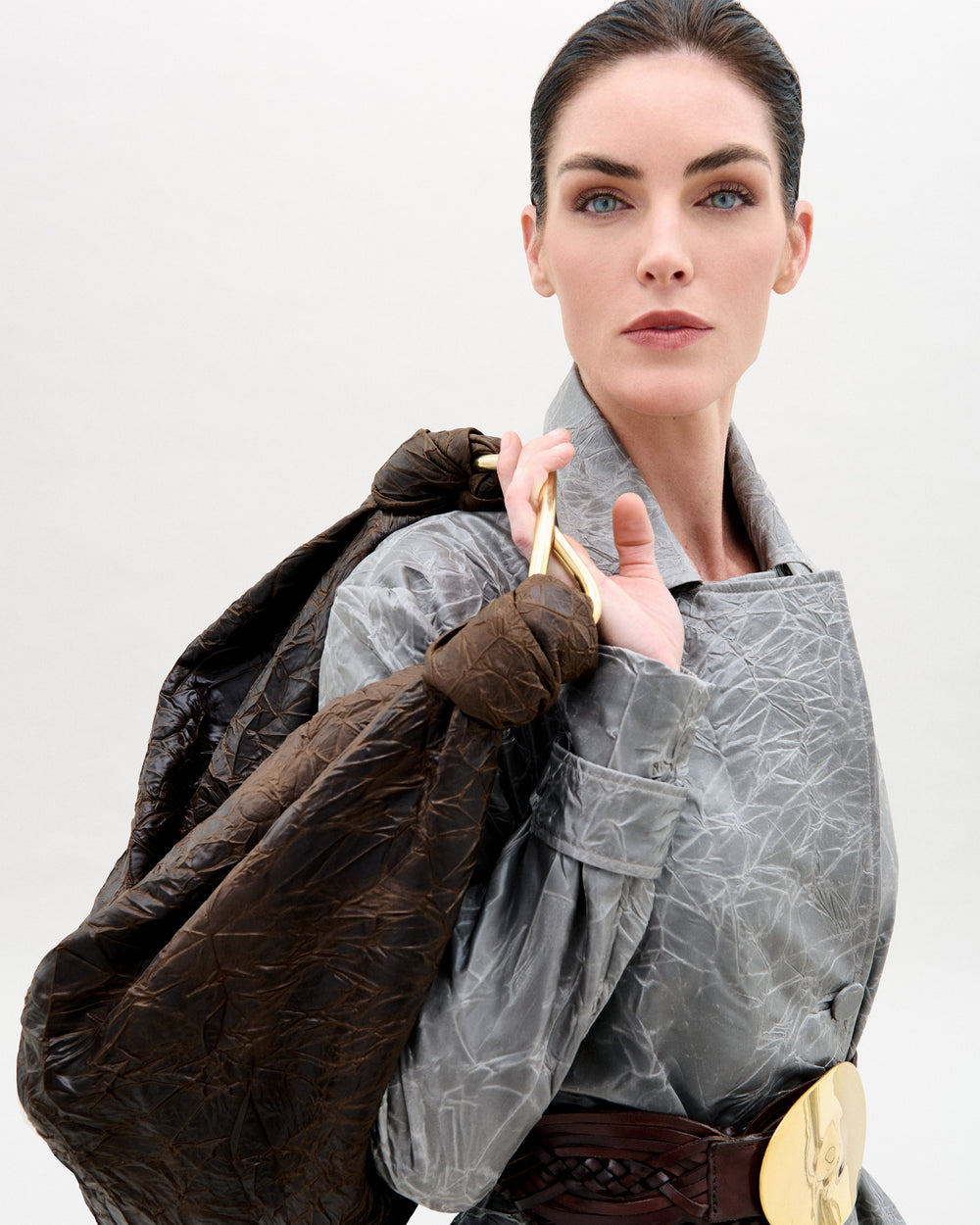 A woman with pale skin and dark hair pulled back wears a textured silver-gray coat, a wide brown belt with a large gold buckle, and carries the Sofia Bag (Maxi) by SVT DESIGNS featuring an S-shaped gold-toned handle over her shoulder.