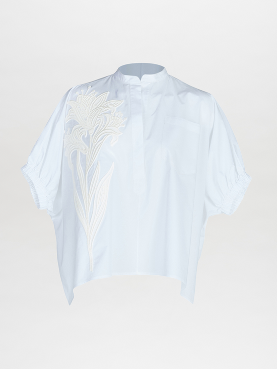 The Silvia Tcherassi Susanne Blouse is a light blue, oversize short-sleeve blouse with a mandarin collar, large white floral embroidery on one side, gathered sleeve edges, and a small chest pocket.