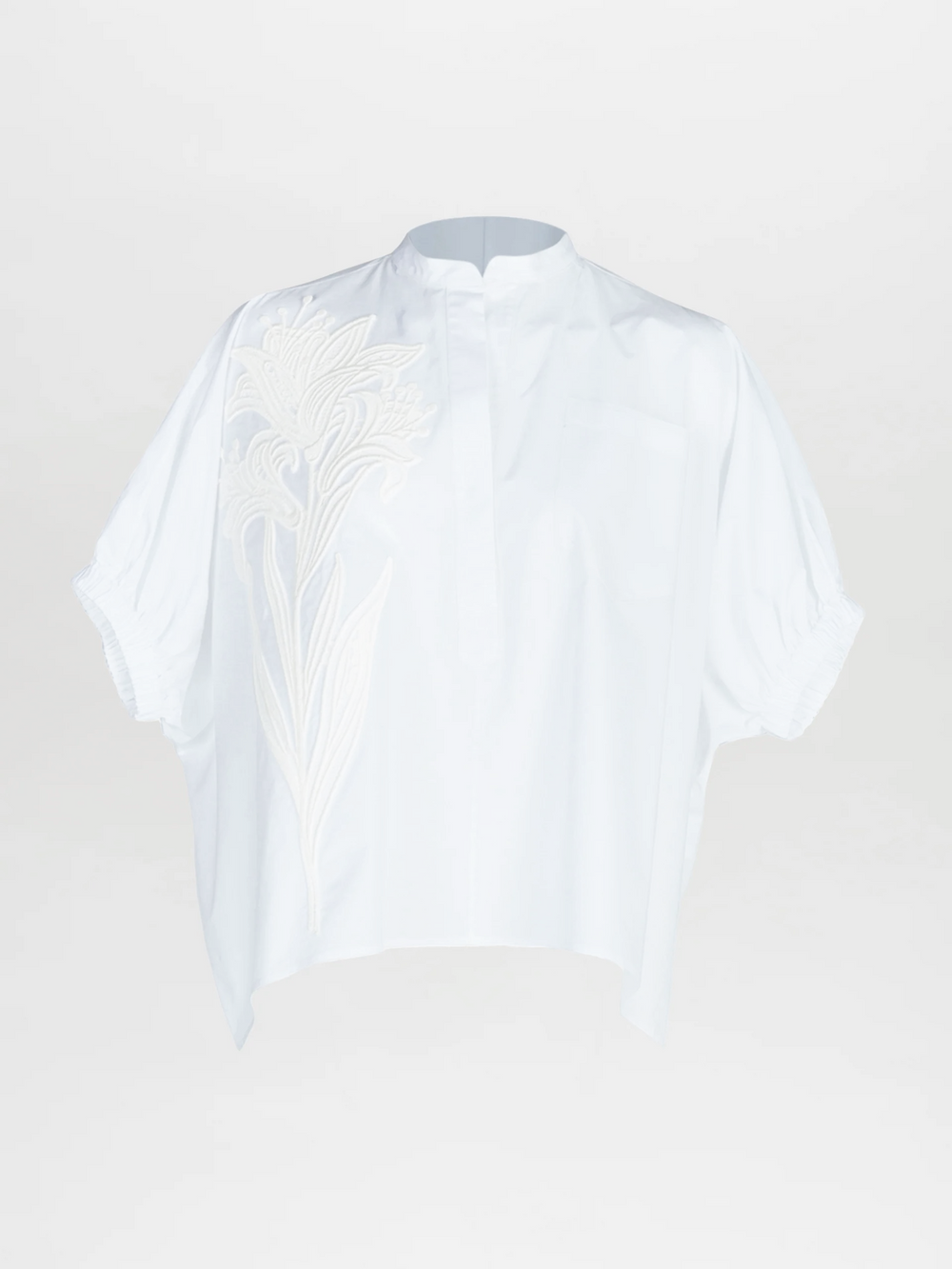 The Susanne Blouse by Silvia Tcherassi is a loose-fitting white top with short, gathered sleeves, a mandarin collar, hidden buttons, and delicate white floral embroidery on the left side.