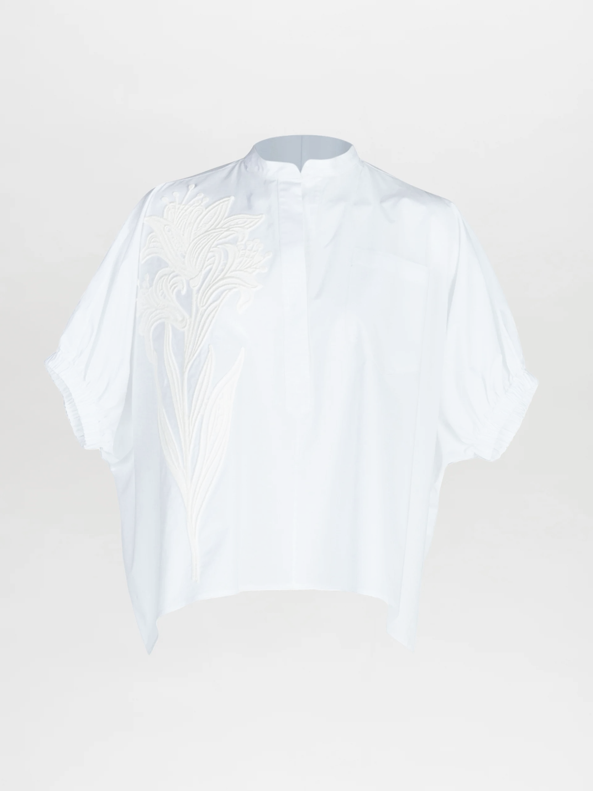 The Susanne Blouse is a loose-fitting, short-sleeved white top featuring a stand collar, subtle button placket, and oversized floral embroidery on the left shoulder and chest.