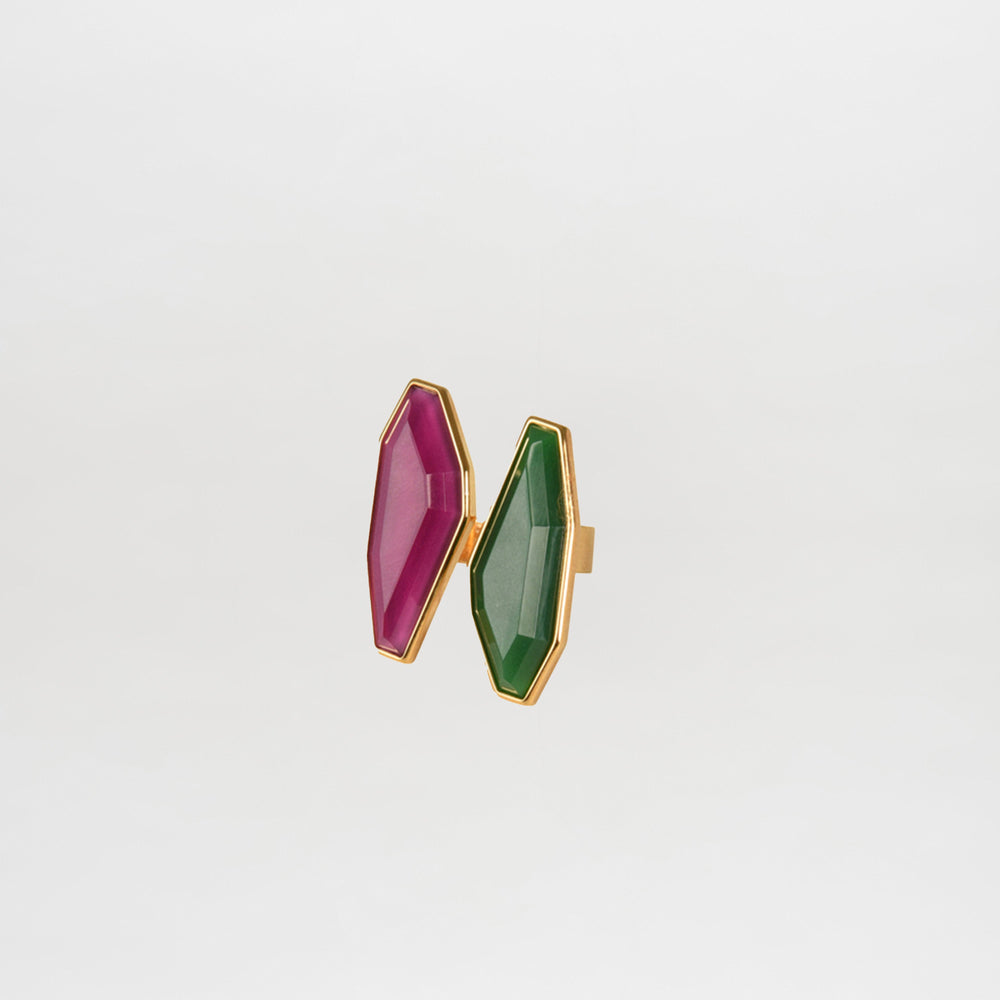The Suzy Ring Magenta Emerald features an asymmetrical gold band with two geometric gemstones—one magenta, one emerald—displayed side by side on a plain white background.