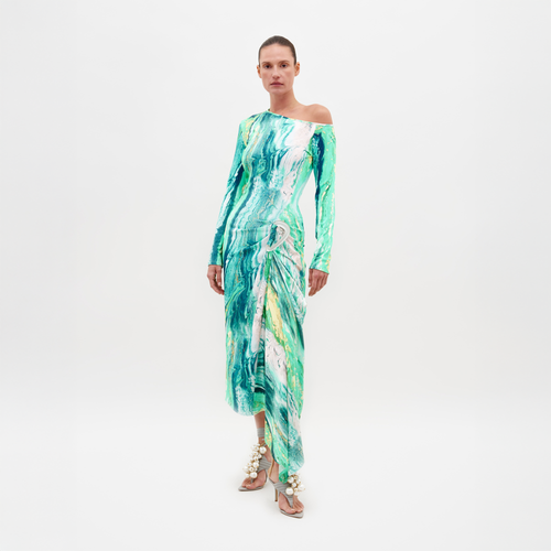 A person stands against a plain white background, wearing the Tahia Dress by SVT DESIGNS, featuring an asymmetric neckline and a wavy green and white abstract print. They are also wearing sandals adorned with large shells.