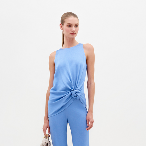 A person with long hair tied back is wearing the SVT DESIGNS Tanima Blouse, a sleeveless blue top in luxurious hammered satin with a knotted waist detail, paired with matching pants. They hold a small bag against a plain white background.