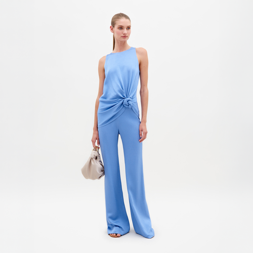 A person poses against a plain backdrop wearing a sleeveless blue top knotted at the waist and SVT DESIGNS's Palermo Pant, which is high-waisted with wide legs made from fine Italian textile. They're carrying a small beige handbag and have long hair tied back.