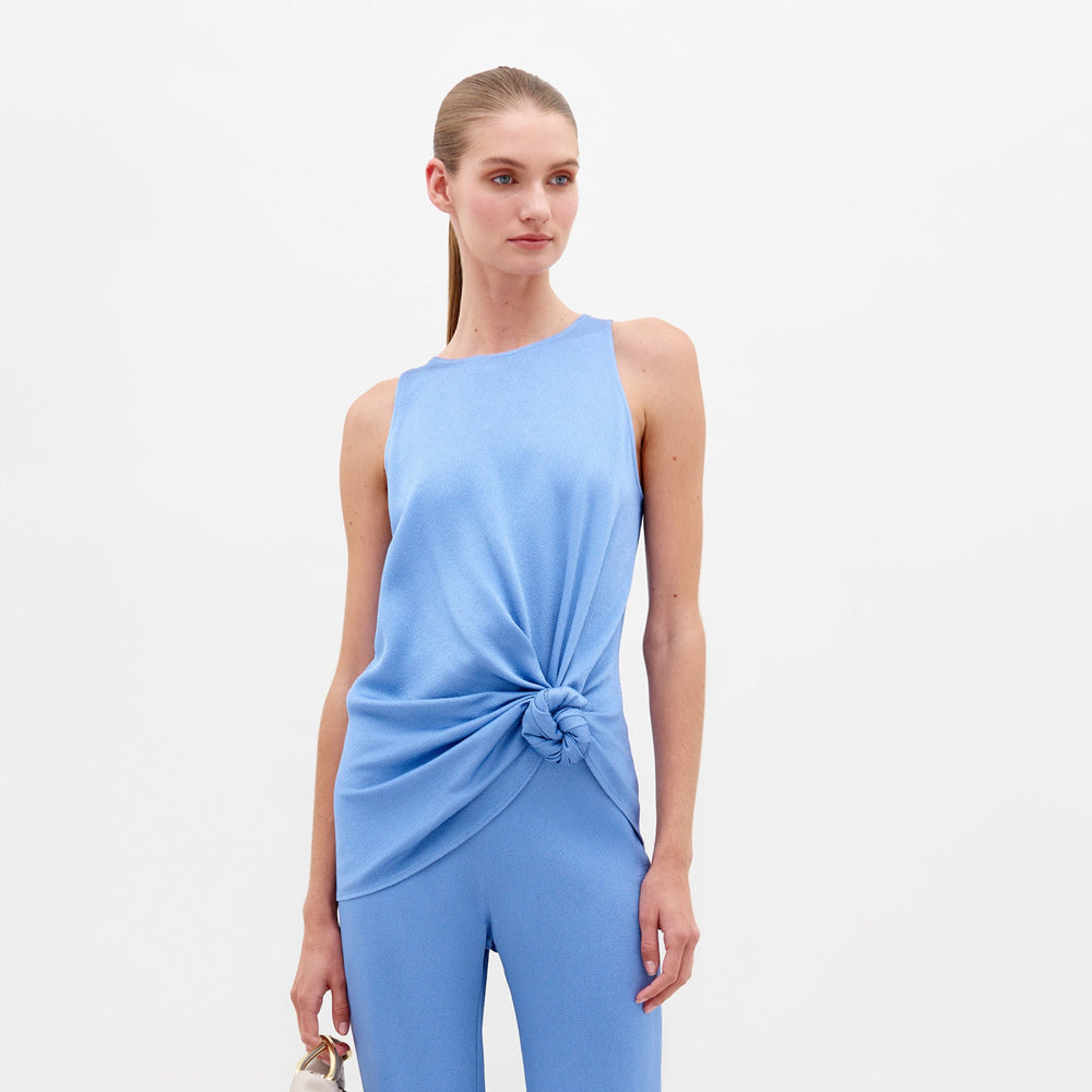 A person wearing the Tanima Blouse in periwinkle, a light blue sleeveless top with a knot at the waist, paired with matching pants and holding a small handbag, exudes effortless style. Perfect for pre-order, this chic ensemble ensures you'll be runway-ready by February 15th.