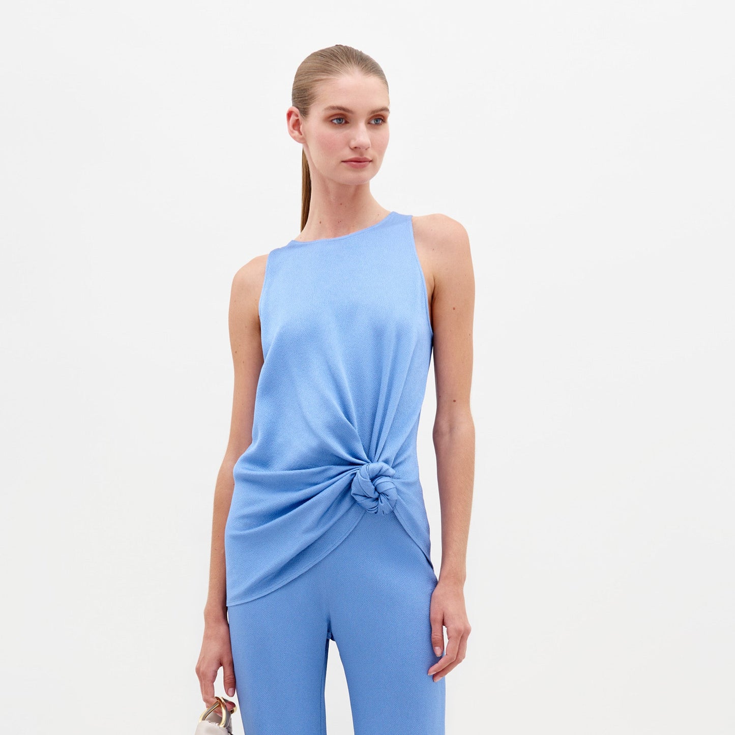 A person wearing the Tanima Blouse in periwinkle, a light blue sleeveless top with a knot at the waist, paired with matching pants and holding a small handbag, exudes effortless style. Perfect for pre-order, this chic ensemble ensures you'll be runway-ready by February 15th.