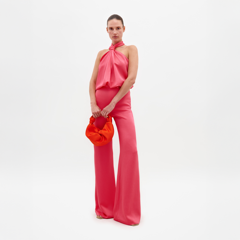 A woman stands against a white background, wearing the Palermo Pant in bright pink with a matching halter-neck top and holding a small round orange bag. She looks at the camera with a neutral expression.