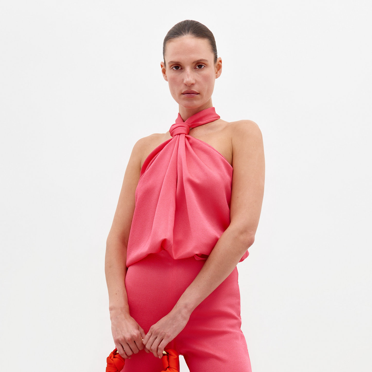 A person stands against a plain background, dressed in the Teagan Blouse Peony—a sleeveless pink top tied at the neck—and matching pink pants, holding a red bag. Available for pre-order now with delivery by February 15th.