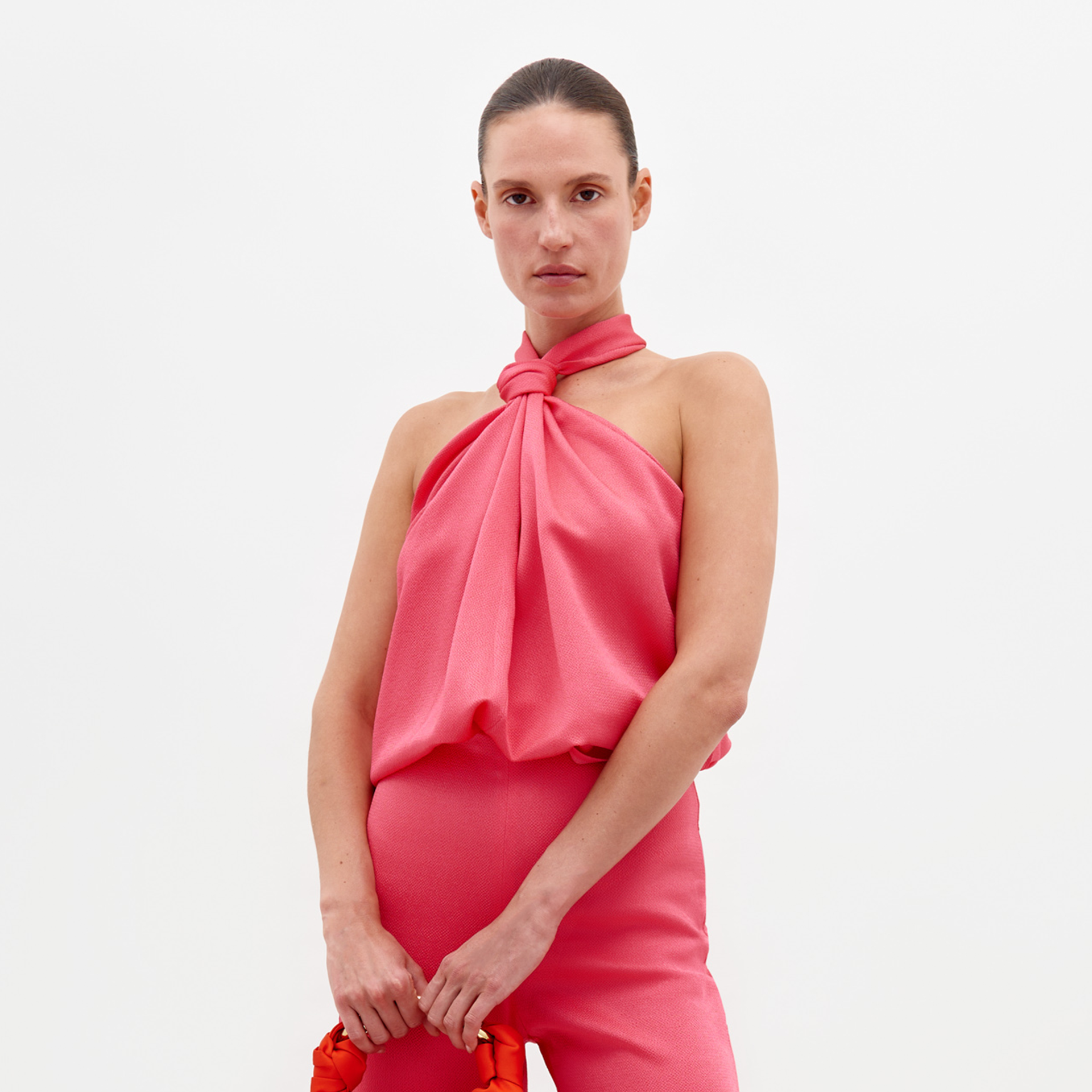 A person stands against a plain background, dressed in the Teagan Blouse Peony—a sleeveless pink top tied at the neck—and matching pink pants, holding a red bag. Available for pre-order now with delivery by February 15th.