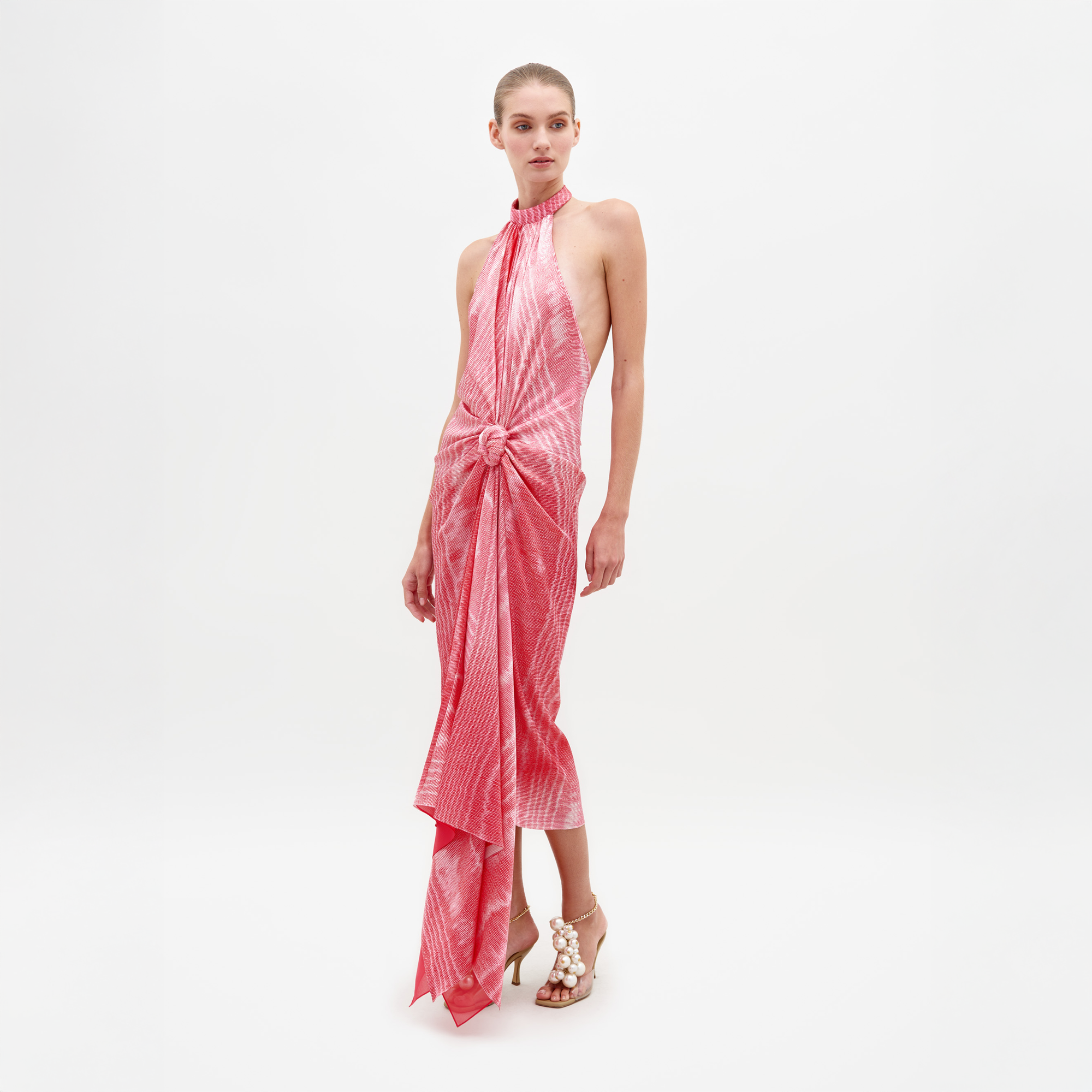 A woman stands against a white background wearing the Theodora Dress by SVT DESIGNS, a sleeveless pink patterned silk midi with a knotted waist and asymmetric hem, paired with open-toe heels adorned with white embellishments.