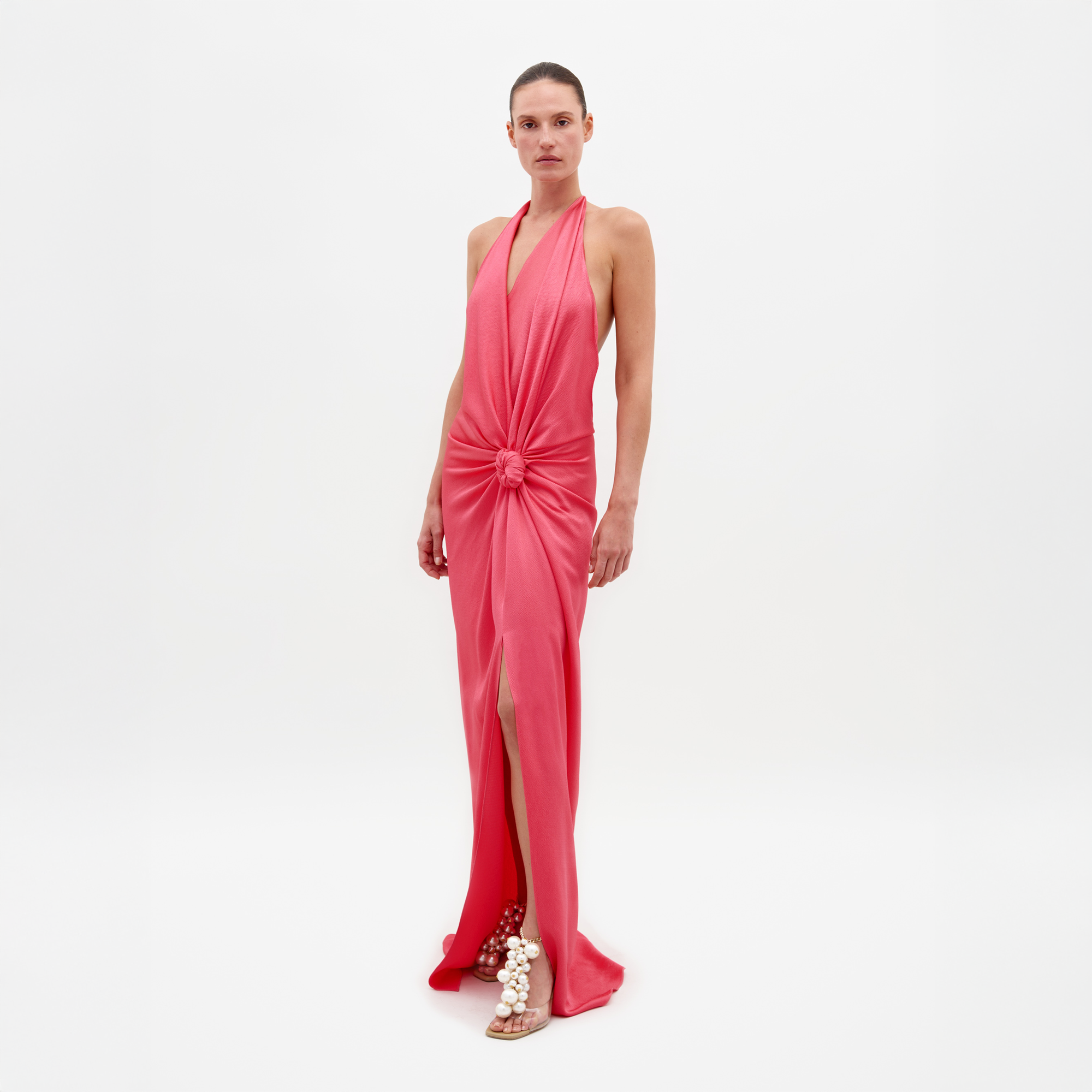 A person stands against a plain white background wearing the SVT DESIGNS Torgiano Dress—a sleeveless, floor-length coral pink maxi with a halter neckline, front knot detail, and high slit—paired with pearl-embellished flat sandals.