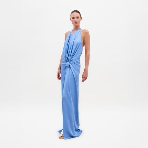 A woman models the SVT DESIGNS "Torgiano Dress," a long, blue maxi dress with a waist knot detail, elegant halter neckline, and flowing design made from fine Italian fabric. The plain white background enhances the focus on her attire.