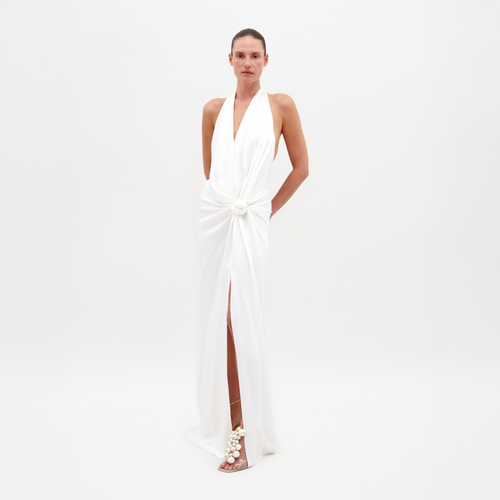 A person wearing the Torgiano Dress by SVT DESIGNS, featuring an Italian textile halter neckline, central knot detail, and side slit, stands against a white background. The dress is paired with sandals decorated with pearl-like embellishments.