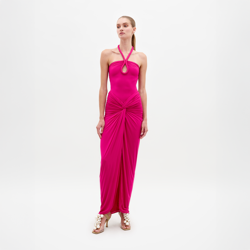 A woman poses against a white background wearing the SVT DESIGNS Tori Dress, a bright pink halter-neck maxi in luxurious Italian fabric with gathered draping at the waist and hem, styled with heeled sandals embellished with large pearls.