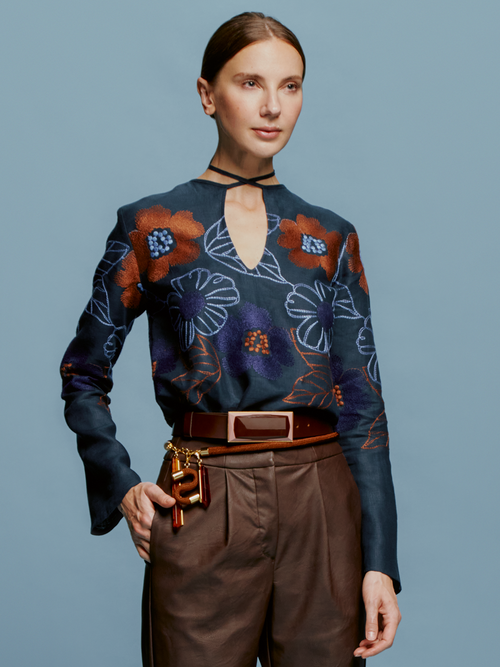 A person stands against a blue background, adorned in the Tosca Blouse Navy Ochre Flower Embroidery with its elegant keyhole neckline, complemented by brown pants and a belt featuring keys and leather accessories. This stylish ensemble could easily be the highlight of fashion week on September 2nd or any day in 2024.