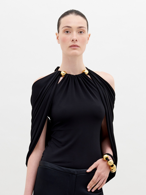 A person with tied-back hair wears a Tabatha Blouse Black with gold accents and a matching gold bracelet, standing against a plain white background. This stylish ensemble is available for pre-order now, with a ship by date of November 15th, 2024.