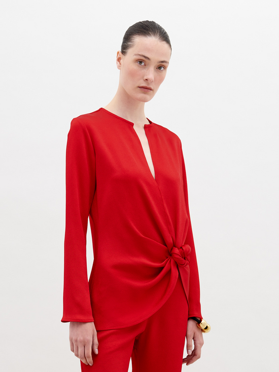 A woman stands in a red outfit with a knot detail on the top. She has her hair pulled back and stares directly into the camera against a plain white background. Available for pre-order now, the Tayra Blouse Rouge will have a ship date of November 15th, 2024.