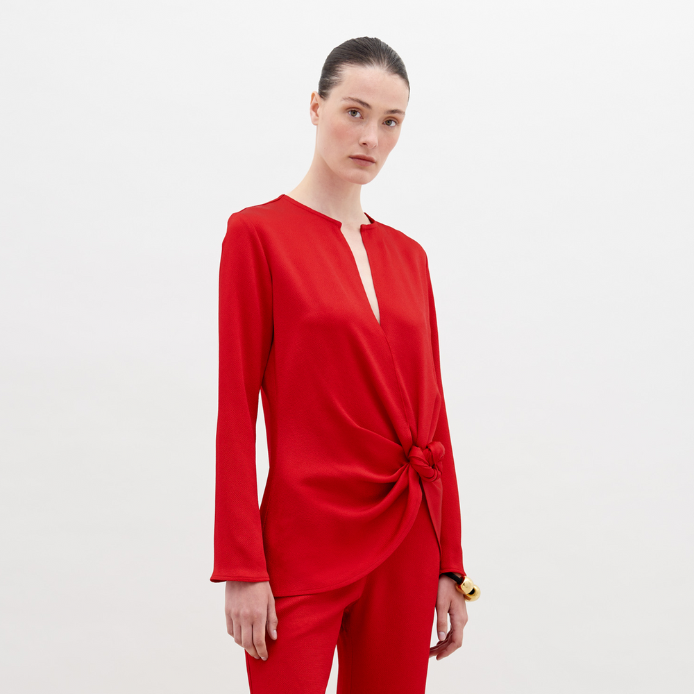 A woman stands in a red outfit with a knot detail on the top. She has her hair pulled back and stares directly into the camera against a plain white background. Available for pre-order now, the Tayra Blouse Rouge will have a ship date of November 15th, 2024.