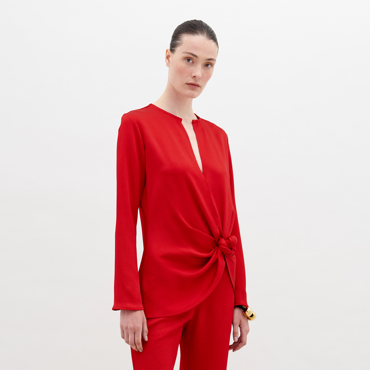 A woman stands in a red outfit with a knot detail on the top. She has her hair pulled back and stares directly into the camera against a plain white background. Available for pre-order now, the Tayra Blouse Rouge will have a ship date of November 15th, 2024.
