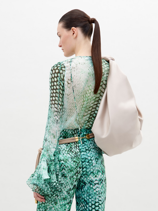 A person with slicked-back hair wears Thea Blouse Emerald Animal Print and holds a large beige bag over their shoulder, perfect for those looking to pre-order now. The background is plain white, highlighting the stylish ensemble expected to ship by November 15th, 2024.