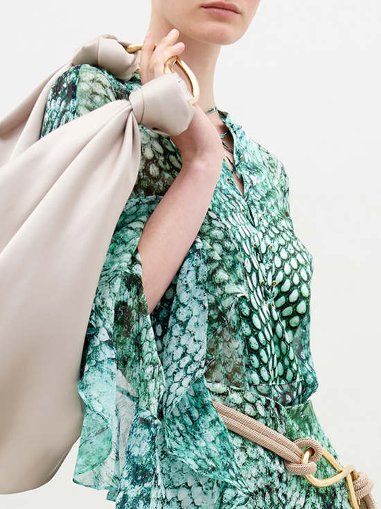 A person with slicked-back hair wears Thea Blouse Emerald Animal Print and holds a large beige bag over their shoulder, perfect for those looking to pre-order now. The background is plain white, highlighting the stylish ensemble expected to ship by November 15th, 2024.