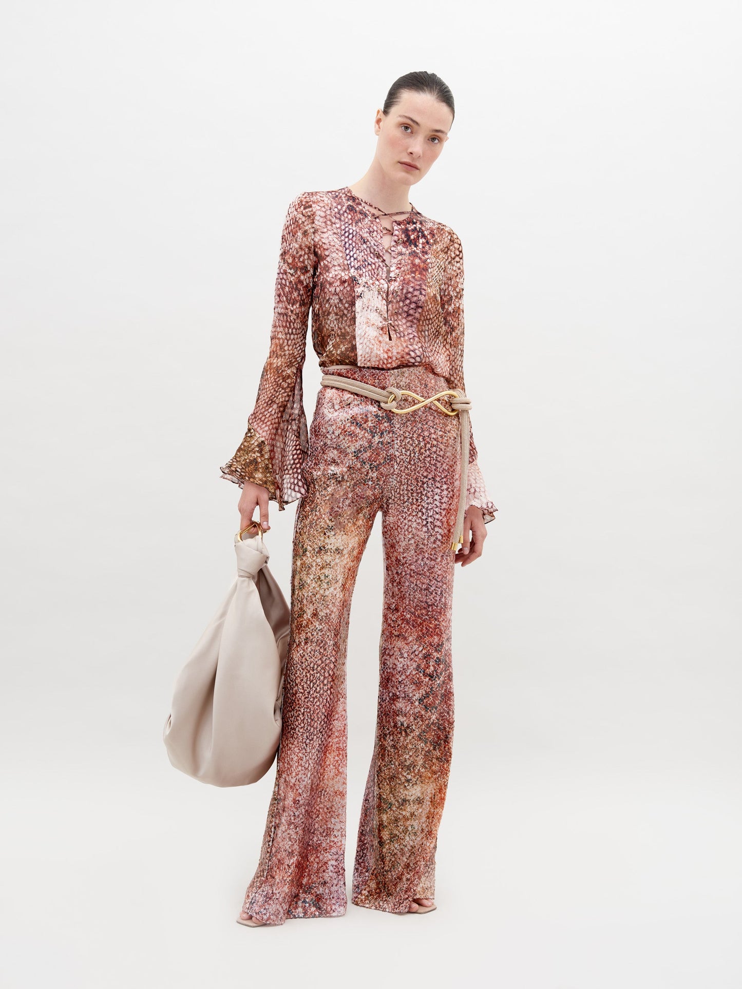 Person wearing a long-sleeved, patterned top and matching Avellino Pant Peach Animal Print with a cinched waist, holding a large beige bag in a white studio setting. Pre-order now to ensure delivery by the ship by date of November 15th, 2024.