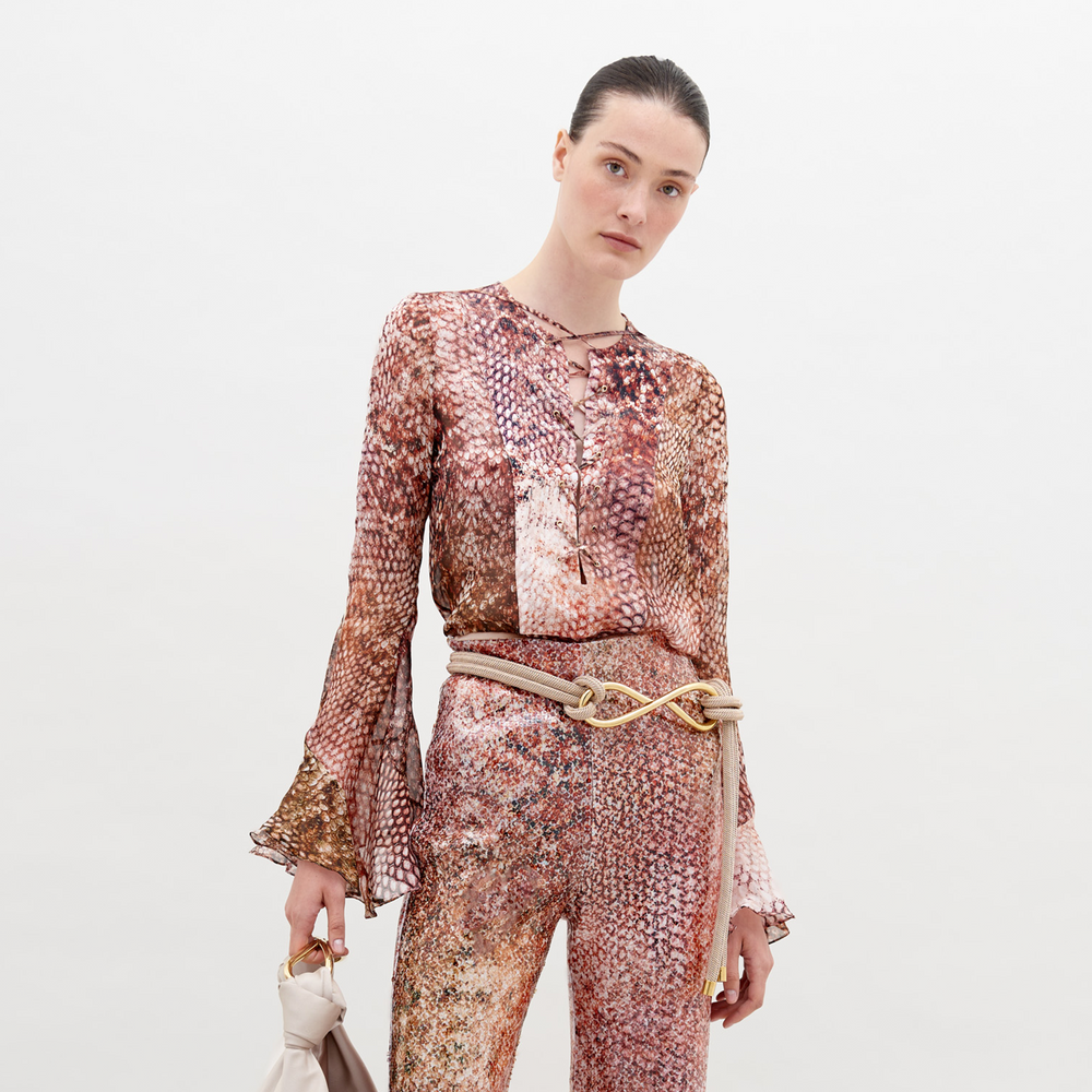 A woman poses against a white background wearing the SVT DESIGNS Thea Blouse, a matching long-sleeve chiffon top and pants in brown and pink snake print, styled with a beige rope belt and a knotted beige bag.