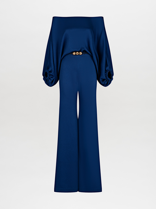 A person stands against a plain background wearing an off-the-shoulder blue top with wide sleeves and matching wide-leg pants, accessorized with a belt that has two large gold embellishments. Pre-order the Theresa Jumpsuit Royal Blue now to ensure you receive your outfit by the ship by date of November 15th, 2024.