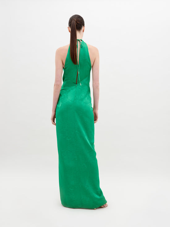 A woman stands against a plain background wearing a sleeveless, high-neck green dress with a front knot and a thigh-high slit. She has her hair pulled back and wears open-toed gold sandals. The Tizziana Dress Green is available for pre-order starting November 15th.