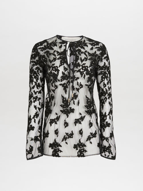 A sheer black and white floral lace blouse with long sleeves and a tie at the neckline elegantly mirrors the Tosca Blouse Black Velvet by Silvia Tcherassi, presented on a plain white background.