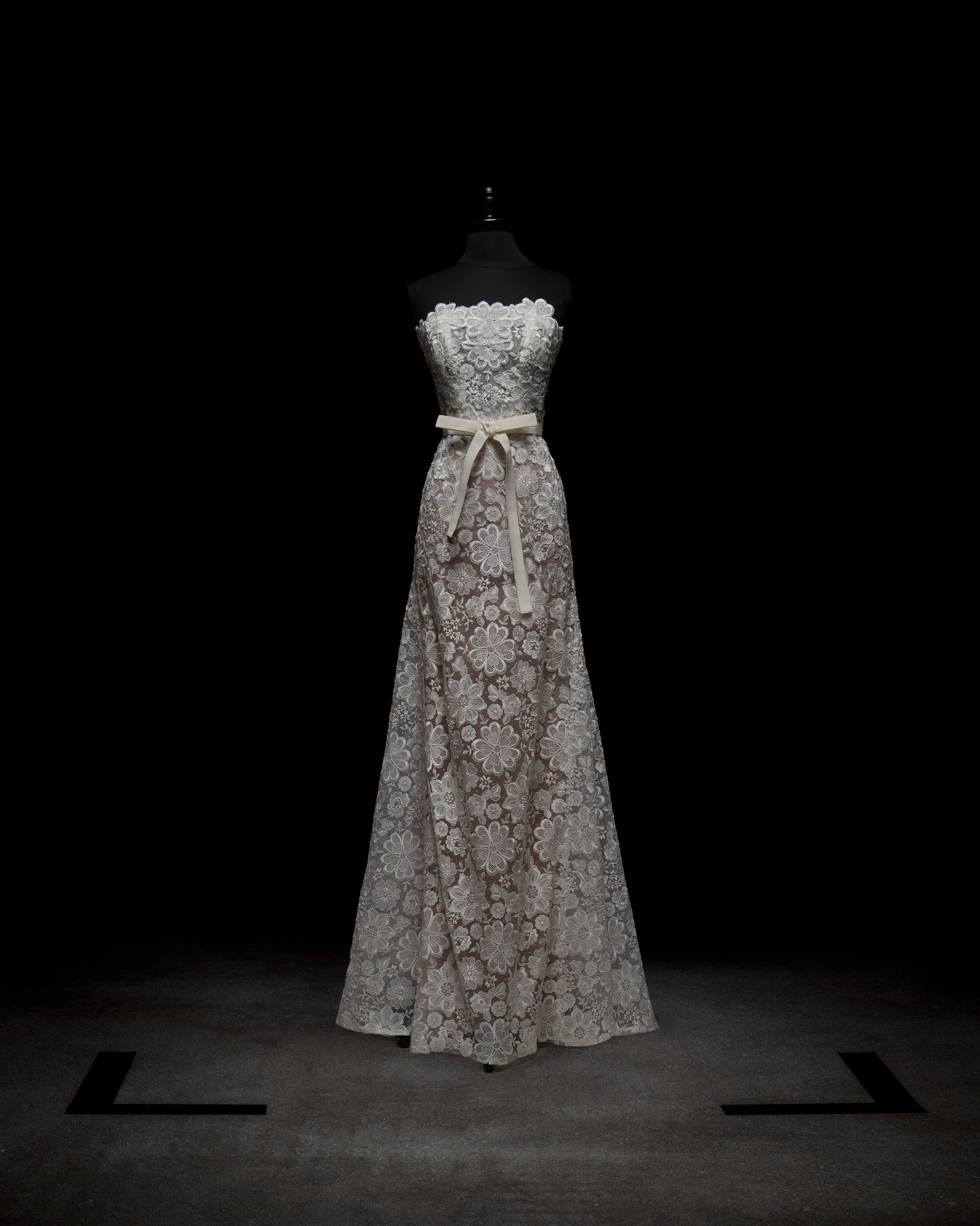 The Valencia Dress by SVT DESIGNS is a strapless, floor-length gown displayed on a mannequin against a dark backdrop. It showcases intricate white lace over light fabric and is accented with a beige ribbon at the waist.