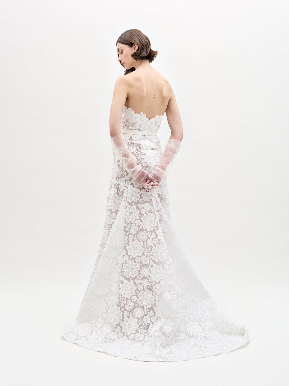 A person in a strapless, floor-length Valencia Dress by SVT DESIGNS stands against a plain white background. Their back is turned, showing short hair and sheer gloves covering hands clasped behind them.
