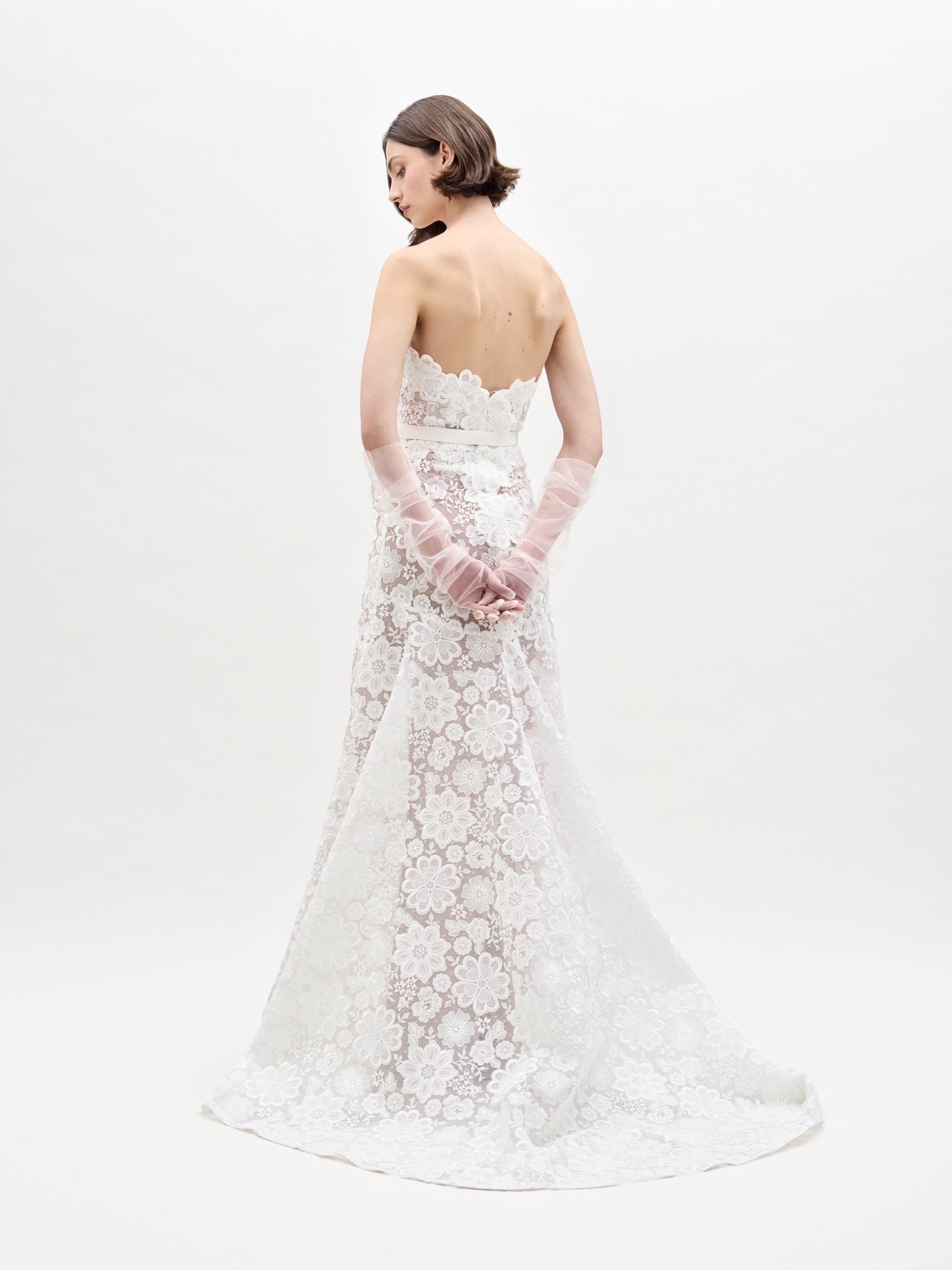 A person in a strapless, floor-length Valencia Dress by SVT DESIGNS stands against a plain white background. Their back is turned, showing short hair and sheer gloves covering hands clasped behind them.