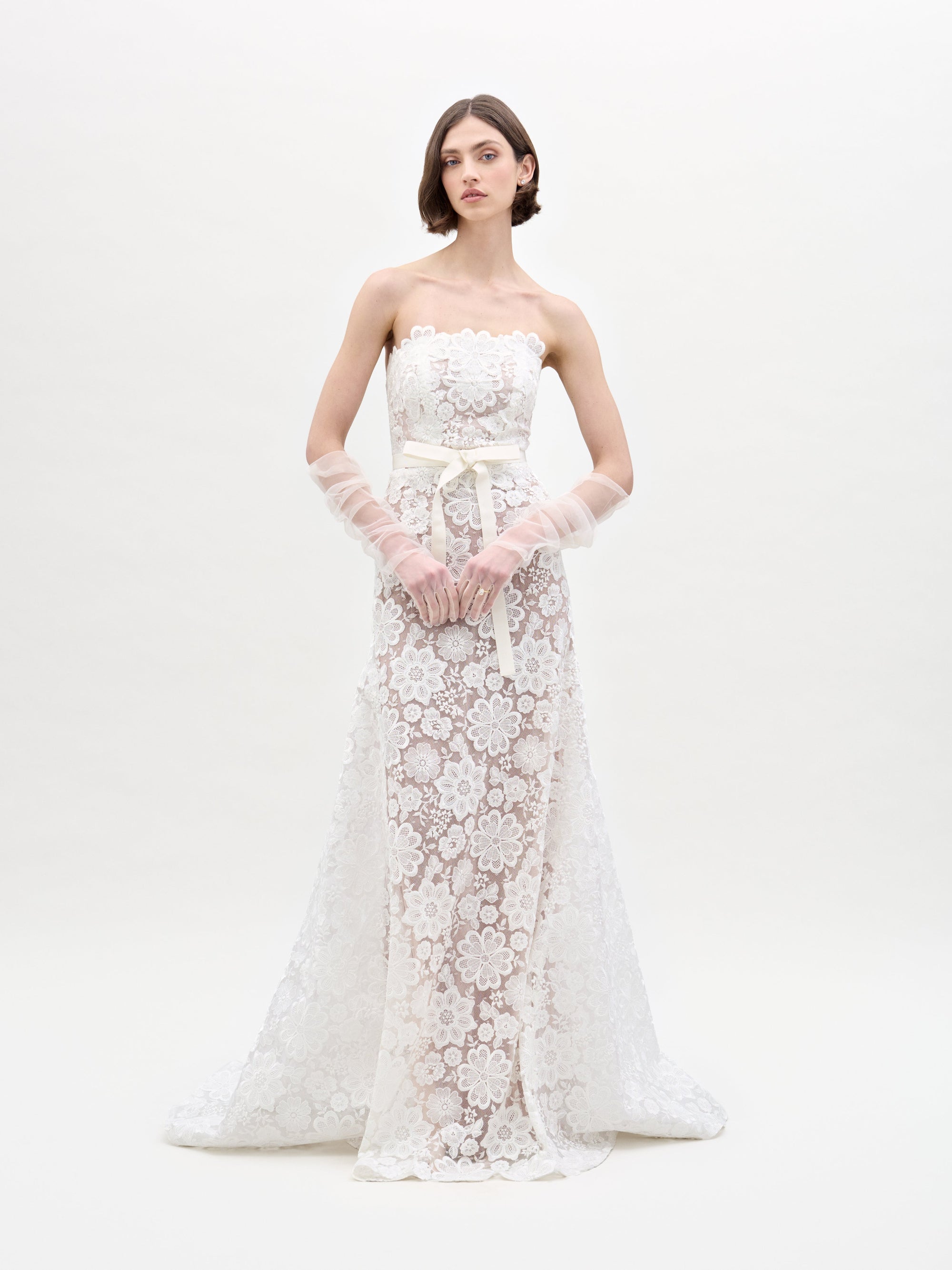 A woman with shoulder-length hair wears the SVT DESIGNS Valencia Dress, a strapless white lace gown featuring floral patterns, a fitted bodice, and a flowing skirt. She pairs it with translucent gloves against an off-white background.