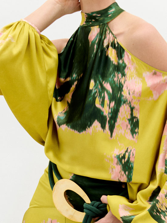 A person wears the Valeria Dress by SVT DESIGNS, a yellow and green patterned maxi dress with exposed shoulders and puffy sleeves. It features pink accents and an abstract design made from fine Italian fabric, cinched at the waist with a wide green belt and large gold buckle against a plain gray backdrop.