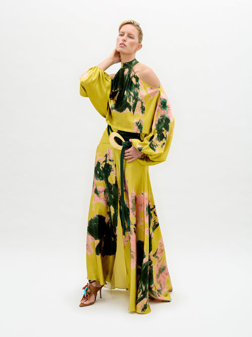 A person poses in the Valeria Dress by SVT DESIGNS against a plain backdrop, showcasing its flowing Italian fabric with yellow, green, and pink patterns. The dress has a halter neckline and exposed shoulders. They pair it with open-toed heels, while resting one hand on their neck and the other on their hip.