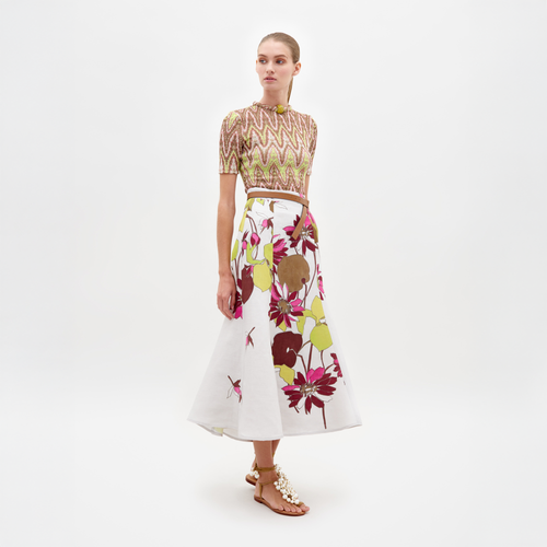 Against a plain white backdrop, someone wears a patterned short-sleeve top with the Erin Skirt by SVT DESIGNS, featuring colorful floral designs in an A-line silhouette. They pair it with embellished sandals and stand gazing forward with arms at their sides.