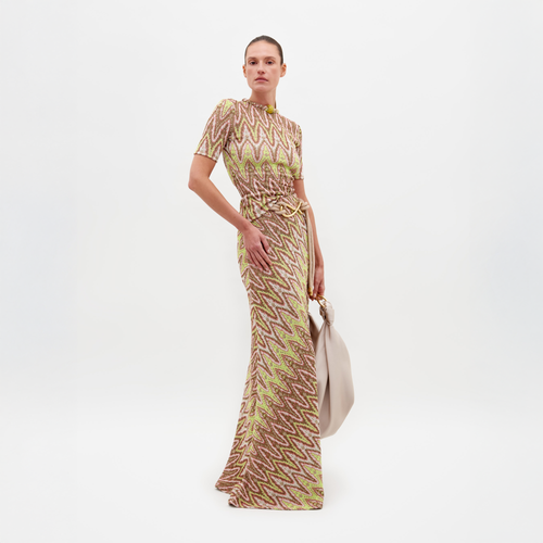 A person models the SVT DESIGNS Famara Skirt with an abstract zigzag pattern in beige, brown, and yellow. They hold a large, light-colored handbag against a plain white background.